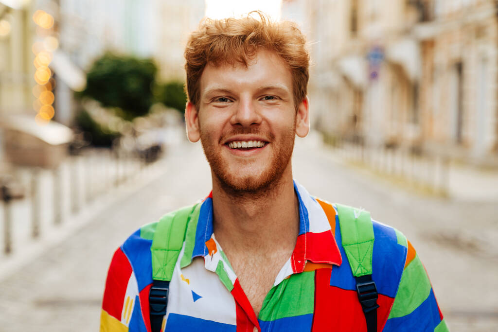 Egy fiatal, jóképű, mosolygós, boldog, vörös hajú férfi portréja, színes ingben, aki a kamerába néz, miközben az utcán áll. - Fotó, kép