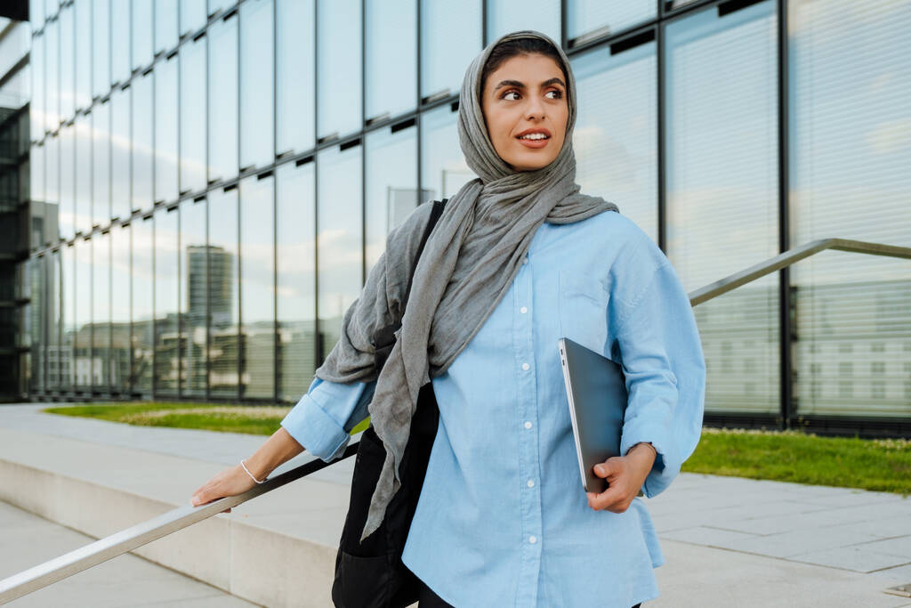 Genç Müslüman kadın dışarıda dizüstü bilgisayarla yürürken başörtüsü takıyor. - Fotoğraf, Görsel