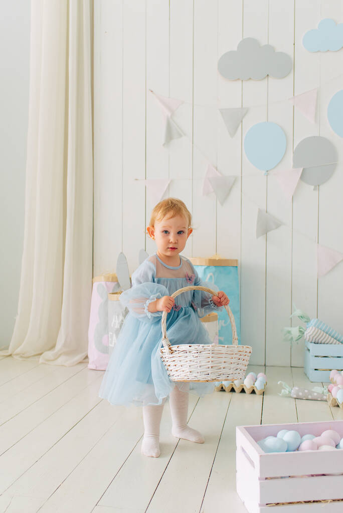 cute little girl w eleganckiej sukience jest trzymając koszyk z dekorowane pisanki. Wielkanoc w urządzonym salonie z dużą ilością pisanek i marchewek dekoracyjnych, selektywne skupienie - Zdjęcie, obraz