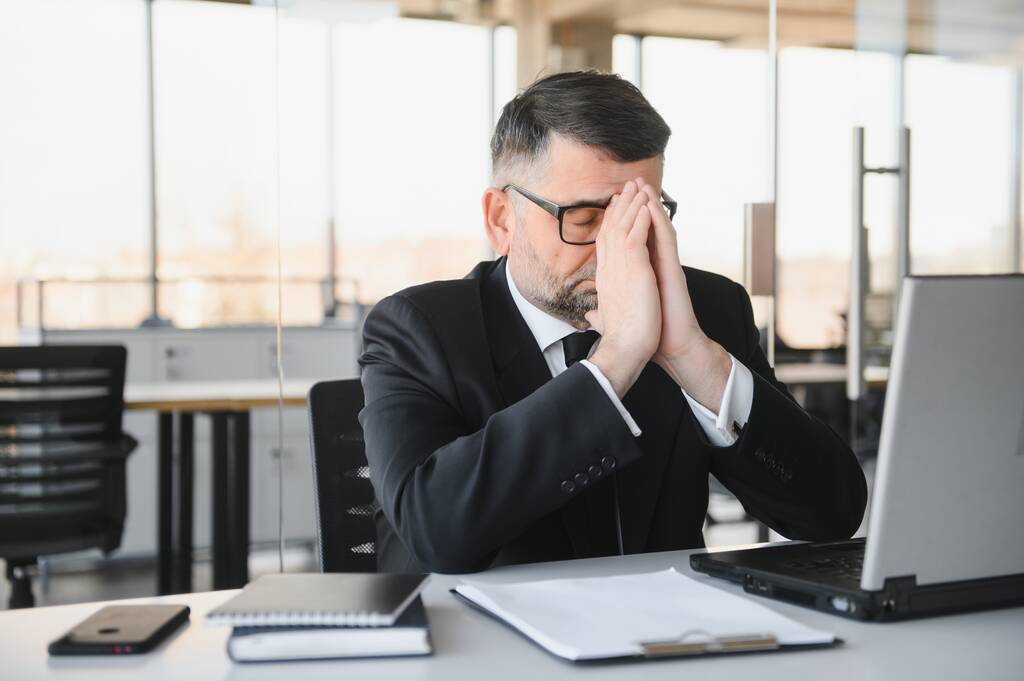 Moe van de gestresste kantoormedewerker die aan zijn bureau zit en nadenkt, wrijft hij over zijn ogen en voelt zich uitgeput - Foto, afbeelding