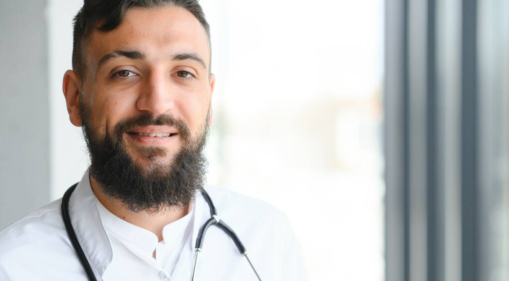 Schöner hispanischer Arzt mit Stethoskop in der Klinik und einem fröhlichen und kühlen Lächeln im Gesicht - Foto, Bild