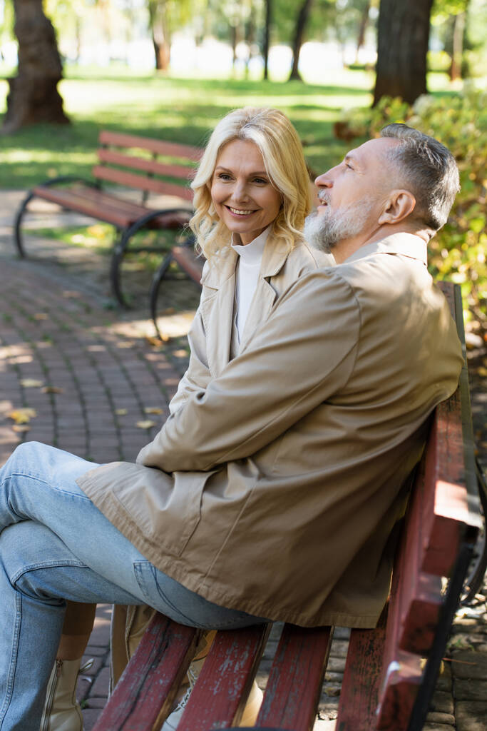 Positive blonde woman sitting near mature husband on bench in park  - Photo, Image