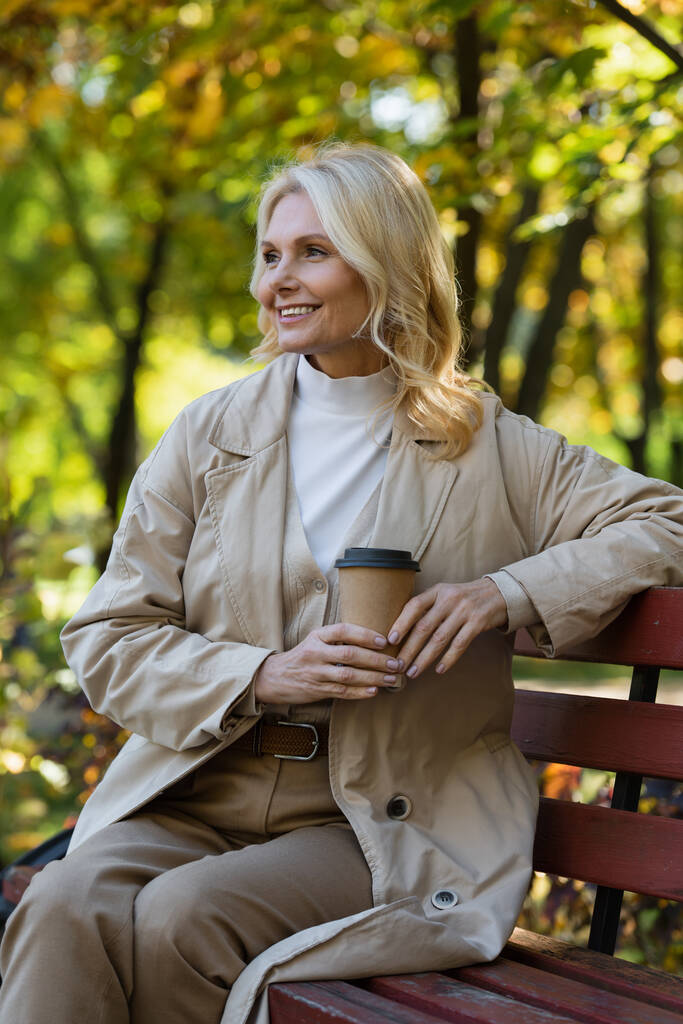 Беззаботная блондинка держит бумажный стаканчик, сидя на скамейке в весеннем парке  - Фото, изображение