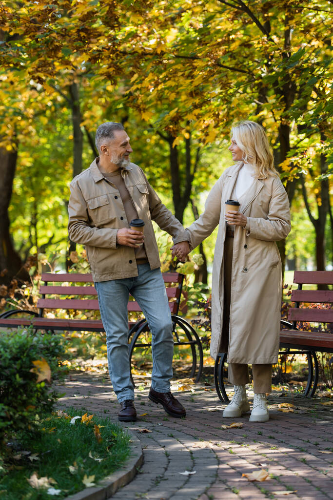 Side view of smiling mature couple holding hands and coffee to go while walking in park  - Photo, Image