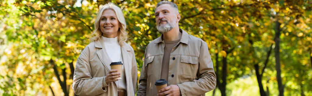 Kaygısız olgun bir çift parkta yürürken kahve içiyorlar, afiş.  - Fotoğraf, Görsel