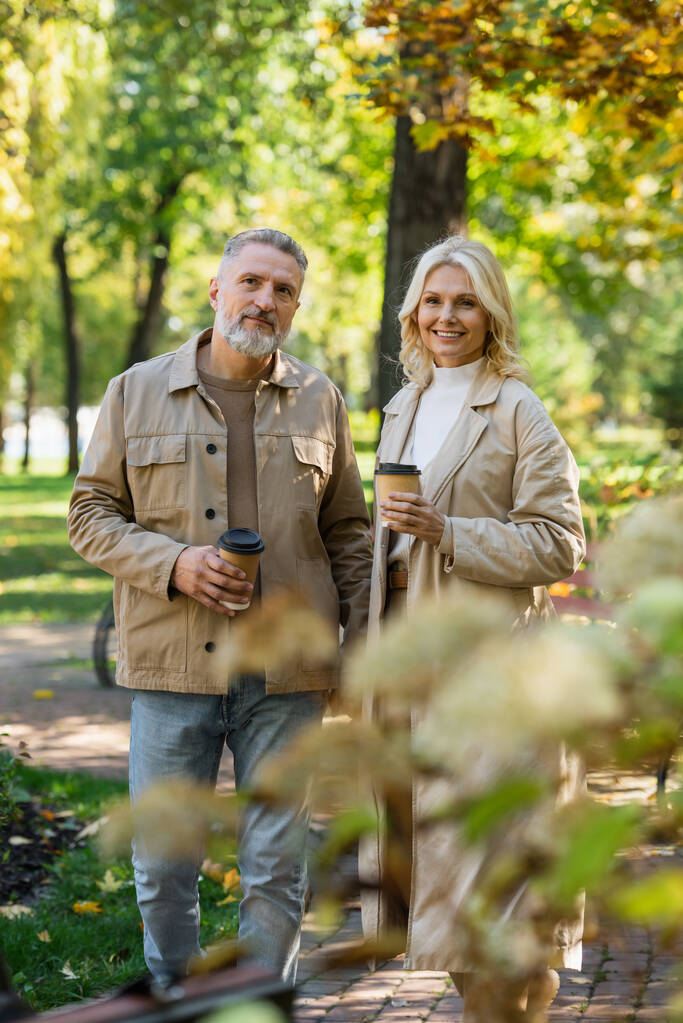 Smiling woman in trench coat holding coffee to go near husband in park  - Photo, Image