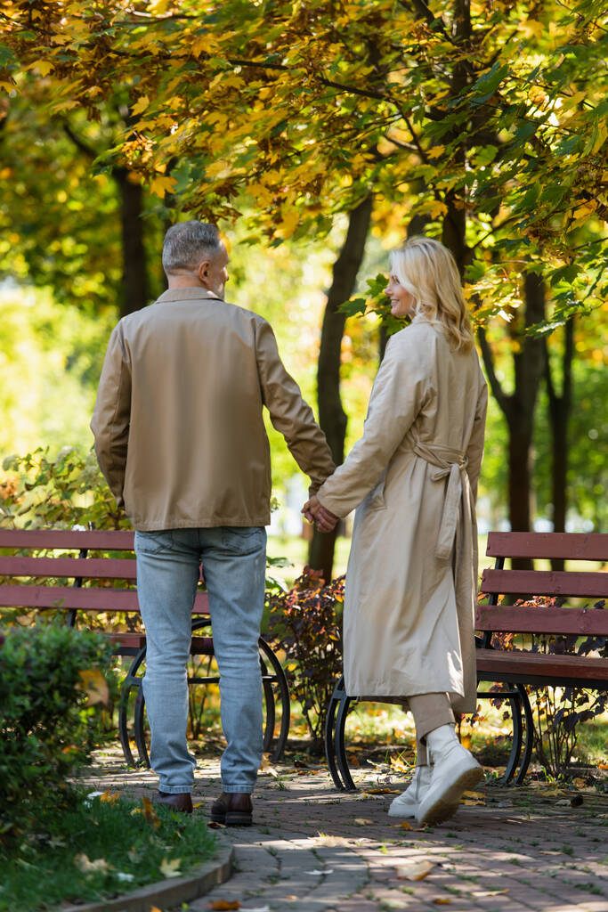 公園を歩いている間に夫の手を握ってトレンチコートでケアフリー女性  - 写真・画像