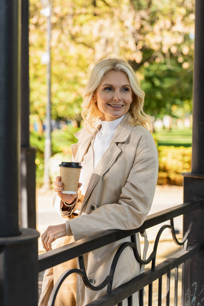 Unbekümmerte Blondine hält Kaffee zum Mitnehmen in der Hand, während sie auf Brücke im Park steht  - Foto, Bild