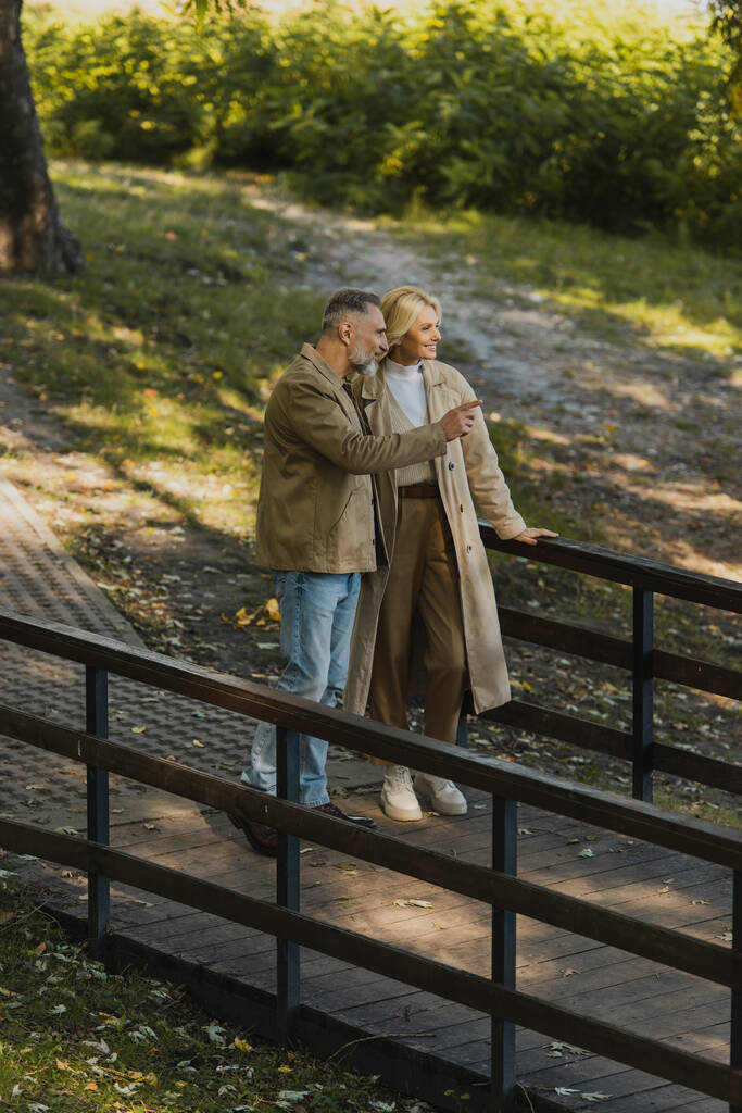 glimlachende volwassen man wijzend met vinger in de buurt van blonde vrouw terwijl staan op de brug in park  - Foto, afbeelding