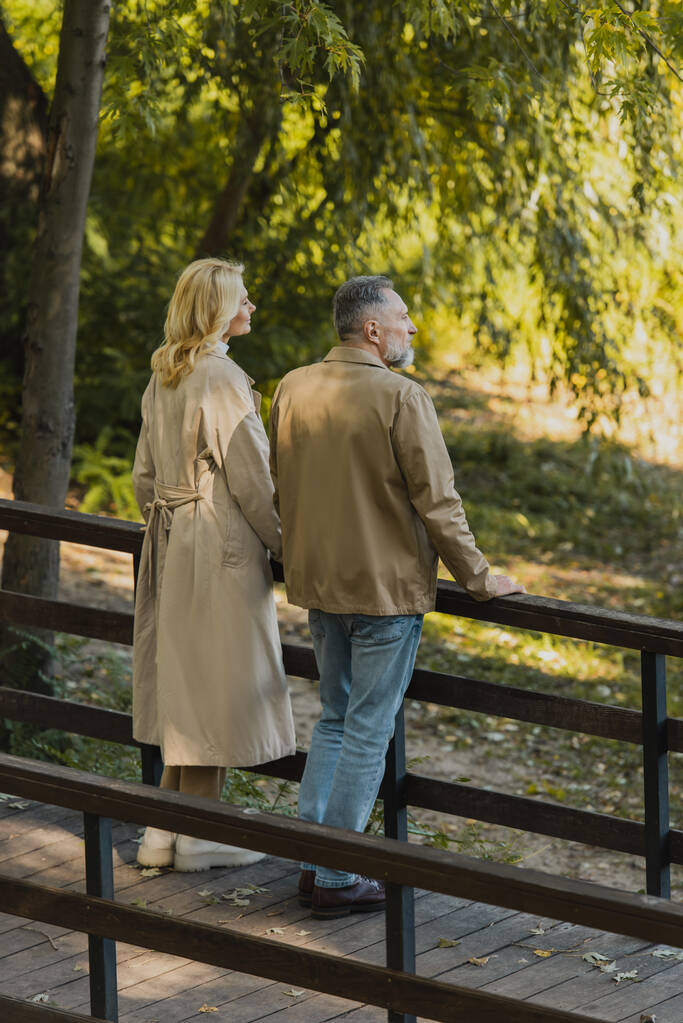 Vista lateral de pareja de mediana edad en traje de primavera de pie en el puente en el parque  - Foto, Imagen