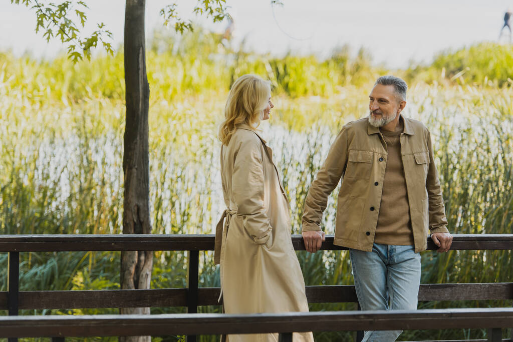 Sorrindo homem maduro olhando para a esposa loira em casaco de trincheira na ponte no parque  - Foto, Imagem