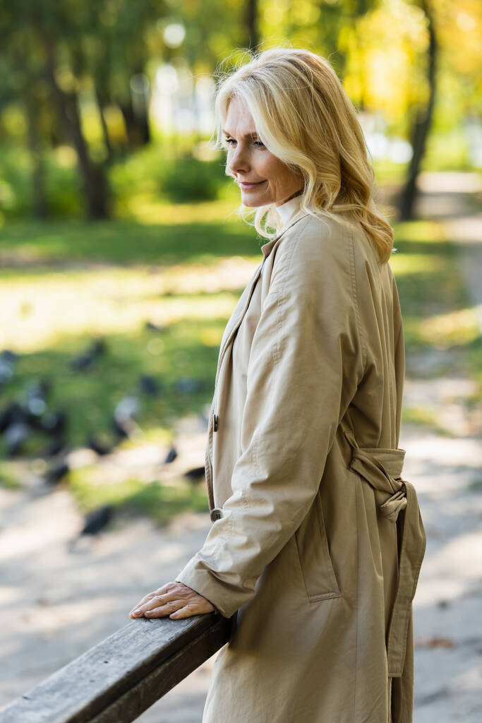 Femme blonde ravie en trench coat debout sur le pont dans le parc de printemps  - Photo, image