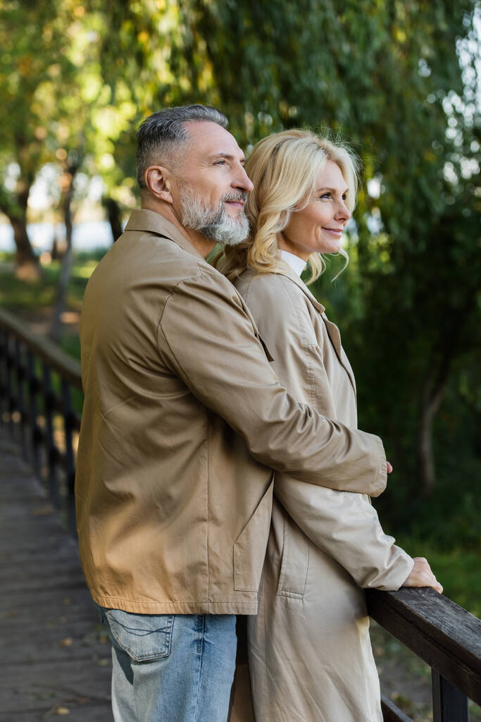 Улыбающийся взрослый мужчина обнимает жену в плаще, стоя на мосту в парке  - Фото, изображение