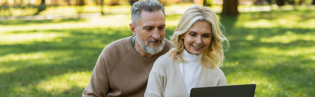 Unbekümmertes Paar mittleren Alters schaut im Frühling Film auf Laptop im Park, Banner  - Foto, Bild