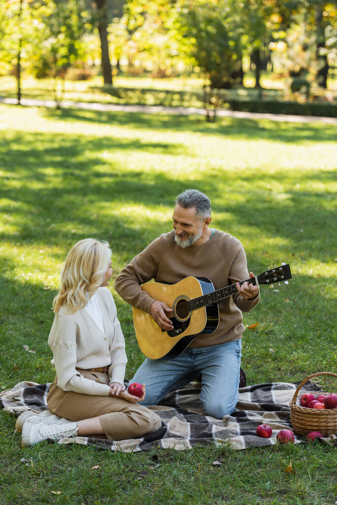 gelukkig man van middelbare leeftijd met grijze baard spelen akoestische gitaar in de buurt van blonde vrouw tijdens picknick in park  - Foto, afbeelding