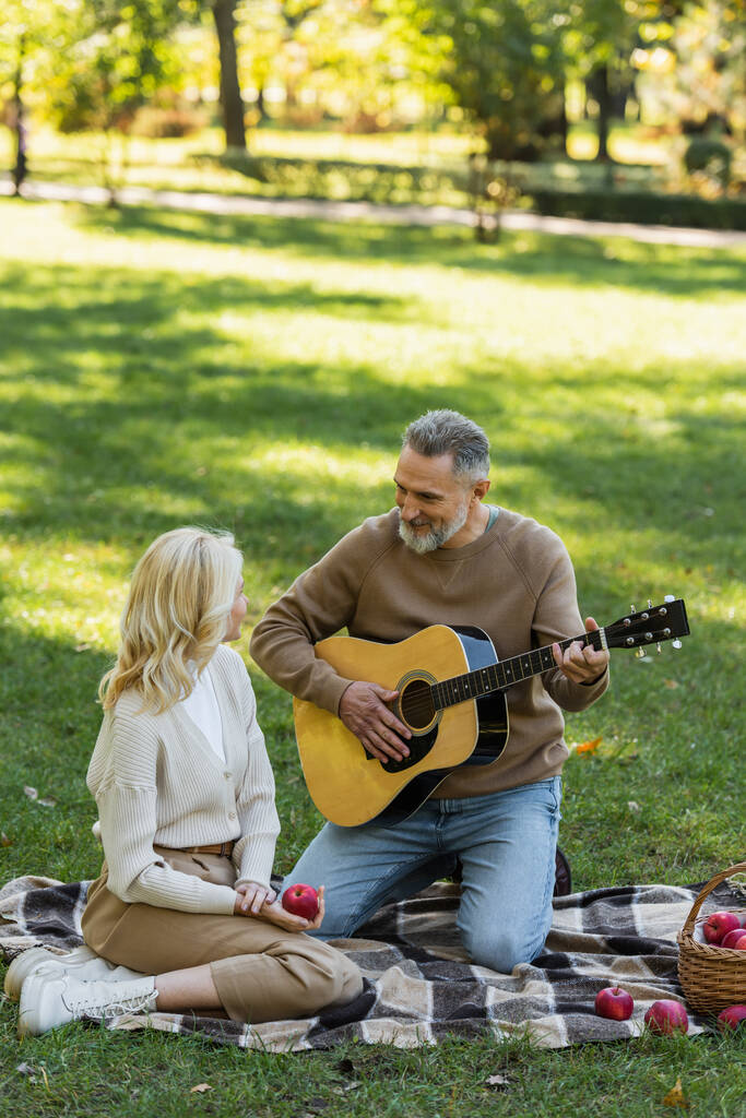 веселый мужчина средних лет с седой бородой играет на акустической гитаре рядом с женой блондинкой во время пикника в парке  - Фото, изображение