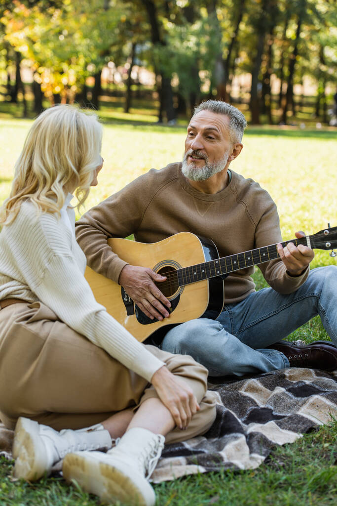 szczęśliwy mężczyzna w średnim wieku z szarą brodą gra na gitarze akustycznej w pobliżu blondynka żona w parku  - Zdjęcie, obraz