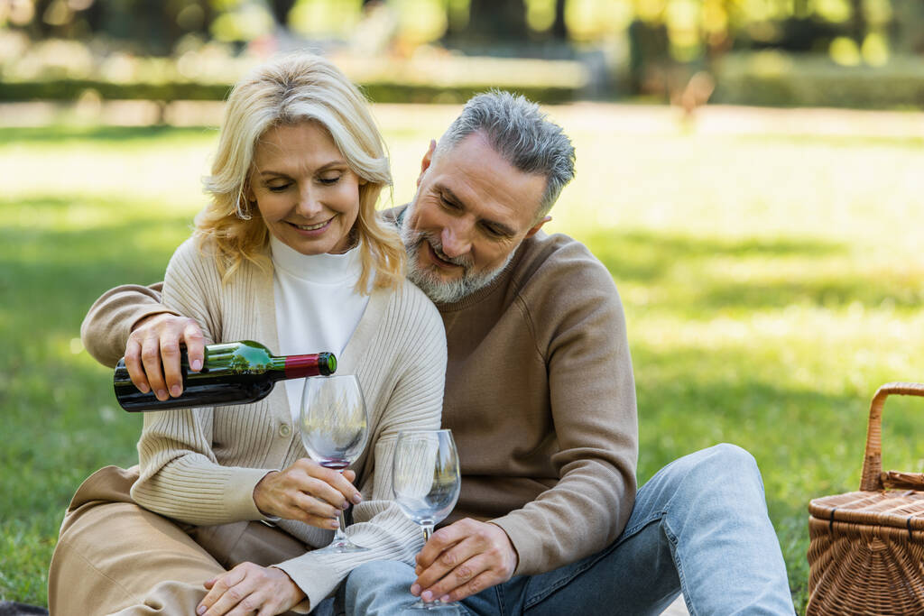 счастливый мужчина средних лет наливает вино в бокал рядом с радостной женой во время пикника в парке  - Фото, изображение
