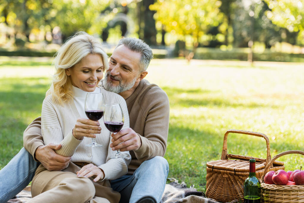 heureux homme d'âge moyen cliquetis verres de vin avec blonde et heureuse femme pendant le pique-nique  - Photo, image