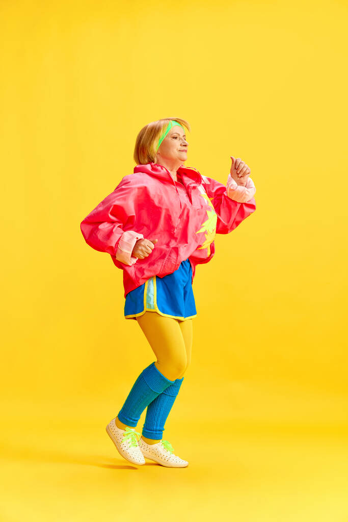 Imagem completa de mulher esportiva idosa em treinamento uniforme colorido, correndo, posando contra fundo de estúdio amarelo. Conceito de estilo de vida esportivo, aposentadoria, cuidados de saúde, bem-estar. Anúncio - Foto, Imagem