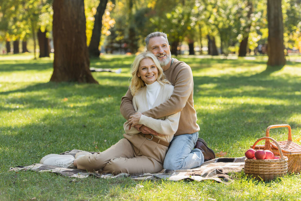 glücklicher Mann mittleren Alters mit grauem Bart umarmt charmante blonde Frau beim Picknick im Park  - Foto, Bild