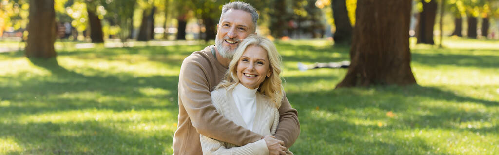 super alegre homem de meia idade com barba cinza abraçando encantadora esposa loira no parque, banner  - Foto, Imagem
