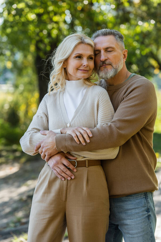 Χαρούμενος μεσήλικας άντρας με κλειστά μάτια αγκαλιάζει ευτυχισμένη σύζυγο στο πάρκο  - Φωτογραφία, εικόνα