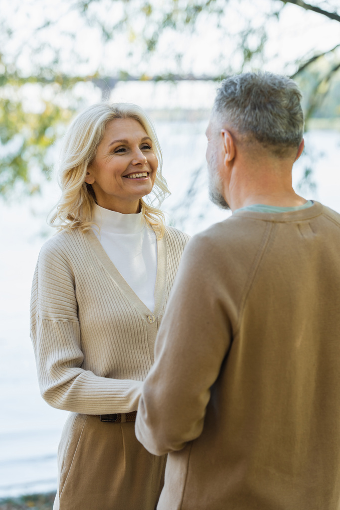 happy middle aged woman looking at bearded husband while holding hands near lake  - Photo, Image