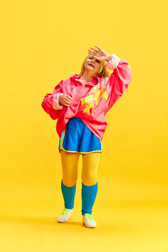 Limpar o suor após o treino. Mulher idosa esportiva em treinamento uniforme colorido, posando contra fundo de estúdio amarelo. Conceito de estilo de vida esportivo, aposentadoria, cuidados de saúde, bem-estar. Anúncio - Foto, Imagem