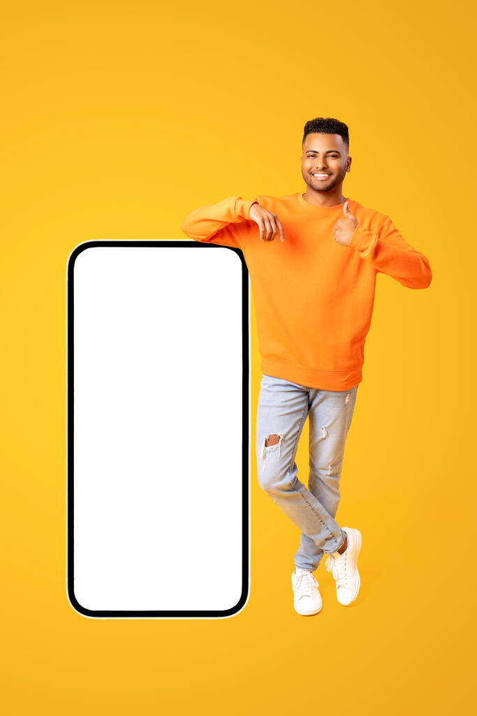 Jóképű fiatal 20-as évek közép-keleti férfi áll teljes hosszában közelében hatalmas okostelefon üres képernyőn, és mutatja hüvelykujj fel, mockup, jóképű indiai srác ajánlott mobil app, kollázs - Fotó, kép