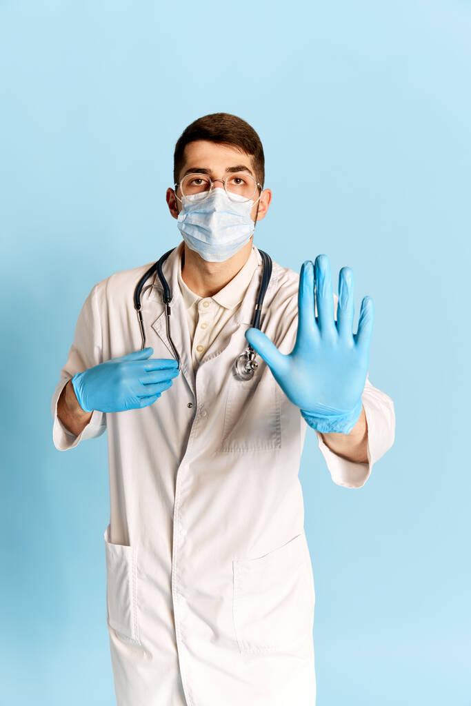 Portrét mladého muže, lékaře v bílých lékařských šatech, ochranné masky a rukavicích pózujících na pozadí modrého studia. Prevence nemocí. Pojem medicína, profese, emoce, povolání. Reklama - Fotografie, Obrázek