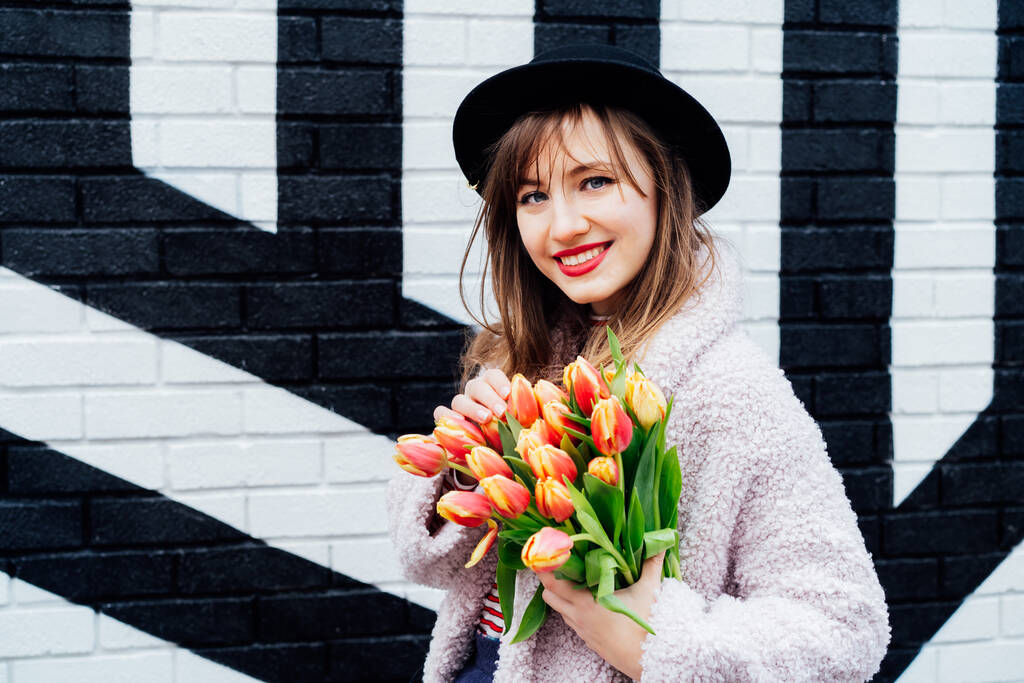 Portret szczęśliwego. uśmiechnięta młoda kobieta moda trzyma bukiet świeżych kwiatów tulipanów na malowanym tle cegły ściany. Miejska moda uliczna. Pozytywny nastrój. Skupienie selektywne. - Zdjęcie, obraz