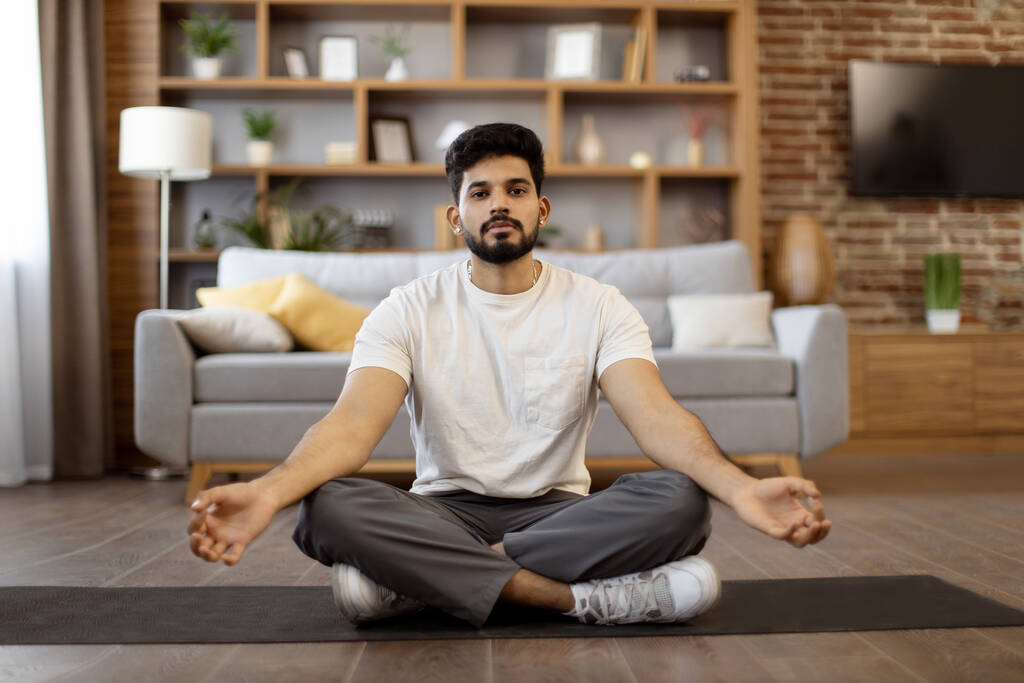 Egészséges, fiatal indiai férfi otthon edz meditációs gyakorlattal. Sportos szakállas fickó ül lótuszpozícióban sárgás gesztussal és a kamerába néz. - Fotó, kép