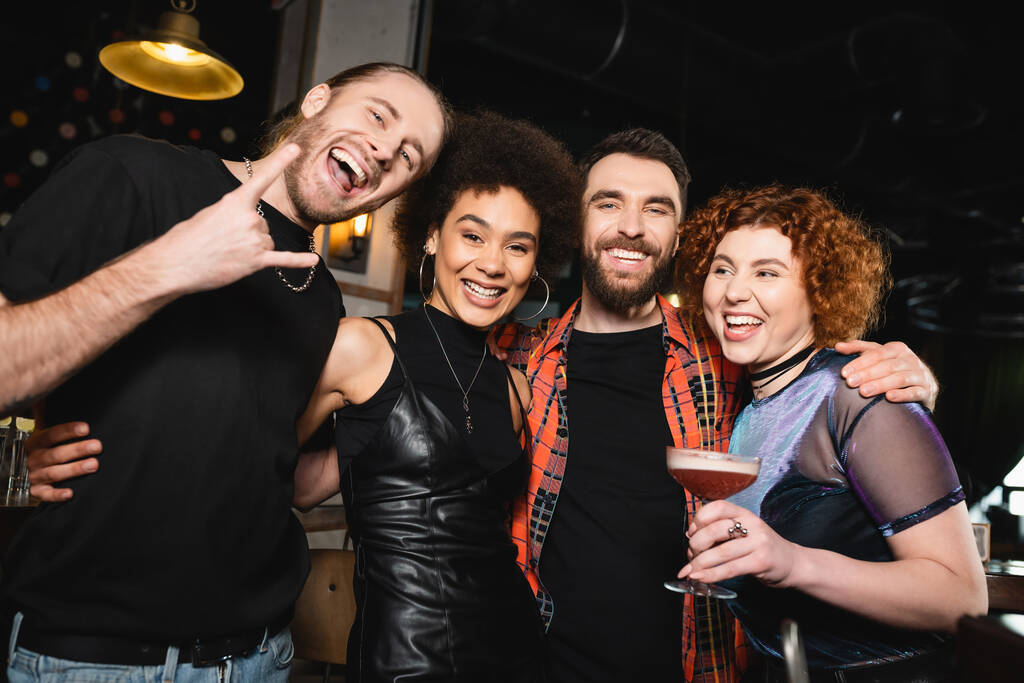 Cheerful redhead woman holding cocktail near multiethnic friends looking at camera in bar  - Photo, Image