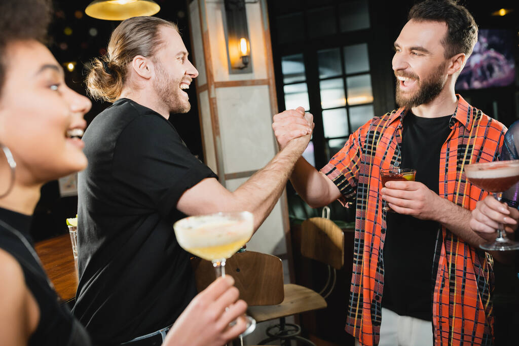 Cheerful bearded friends with cocktails holding hands while meeting in bar  - Photo, Image