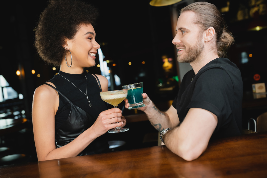 Vista lateral del hombre positivo sosteniendo cóctel de laguna azul cerca de amigo afroamericano en el bar  - Foto, Imagen