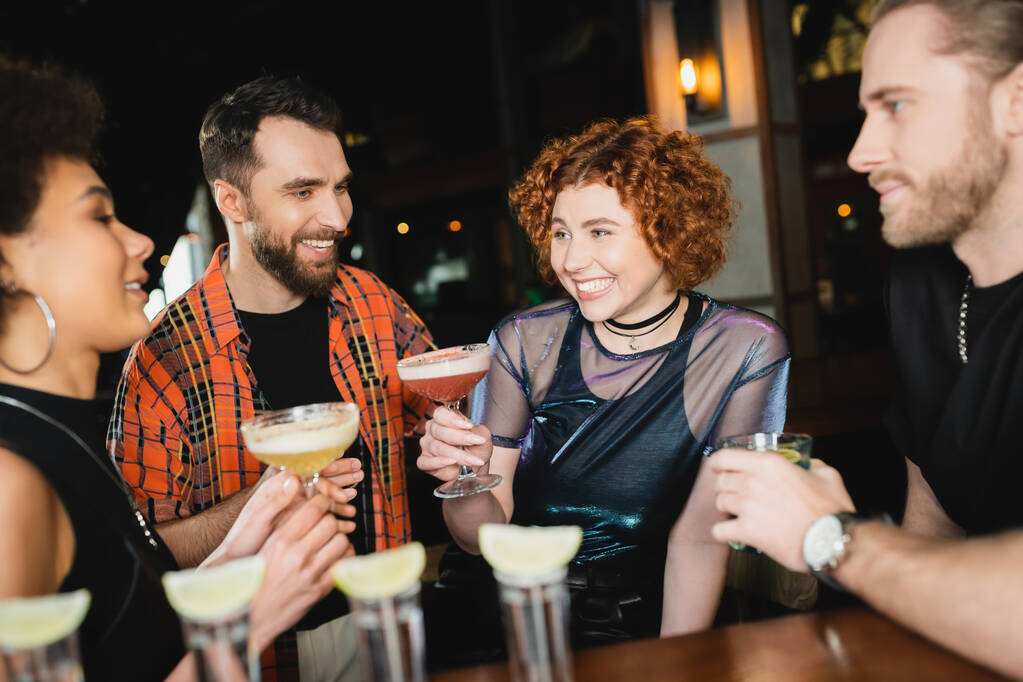 Vrolijke vrouw met cocktail in de buurt van multi-etnische vrienden en tequila shots in de bar  - Foto, afbeelding