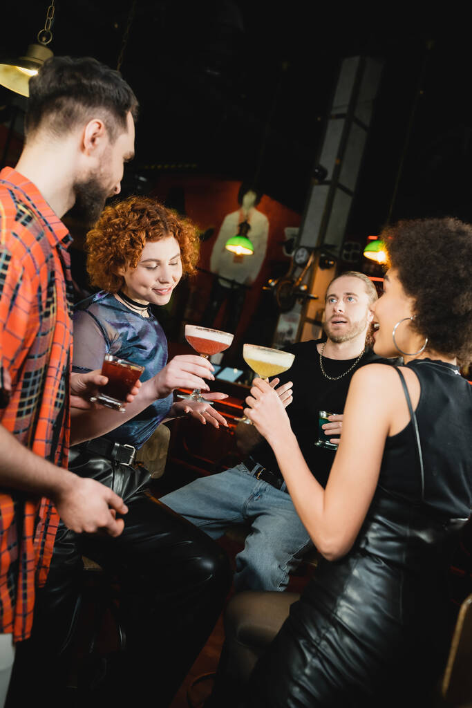 Amigos multiétnicos celebrando diferentes cócteles fríos mientras hablan en el bar  - Foto, imagen
