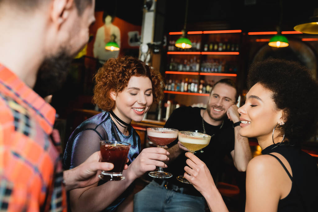 Usmívající se multietnické ženy cinkající koktejly v blízkosti rozmazaných přátel v baru  - Fotografie, Obrázek