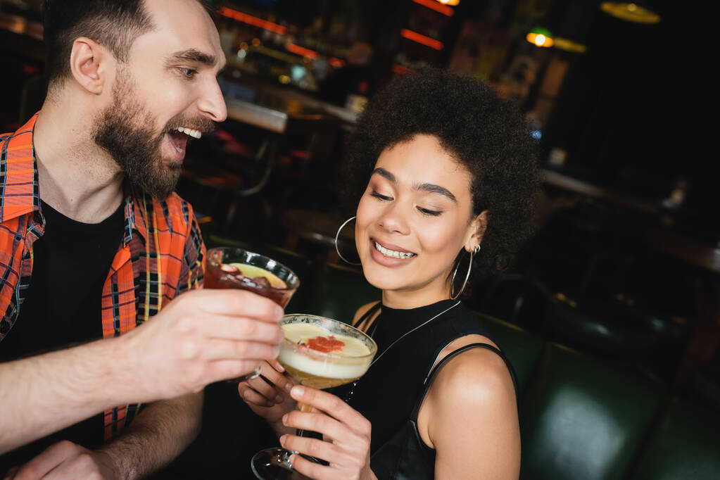 Opgewonden man houdt negroni cocktail in de buurt krullend Afrikaans Amerikaanse vriend in bar  - Foto, afbeelding