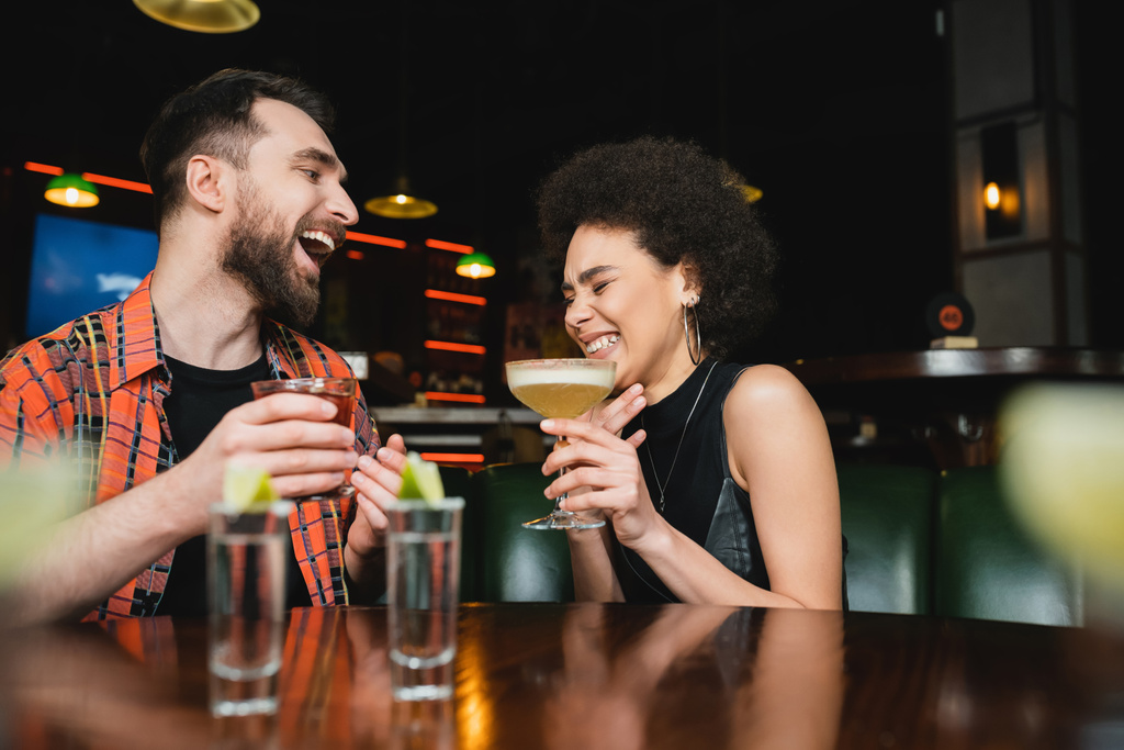 Excited interracial friends holding cocktails and laughing near blurred tequila shots in bar  - 写真・画像