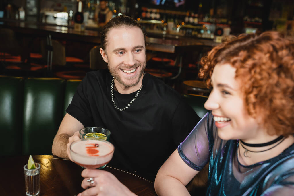 Веселый мужчина с коктейлем из голубой лагуны рядом с размытой рыжей подругой в баре  - Фото, изображение