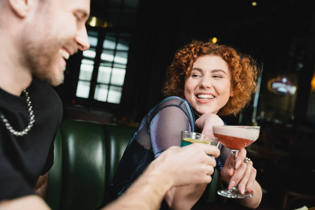 Позитивная рыжая женщина, звенящая коктейлем клуба клевера с размытым другом в баре  - Фото, изображение
