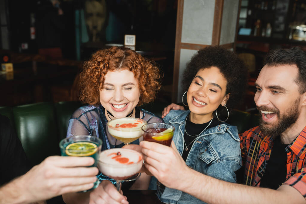 positieve interraciale vrienden glimlachen en toasten met verschillende cocktails in bar  - Foto, afbeelding