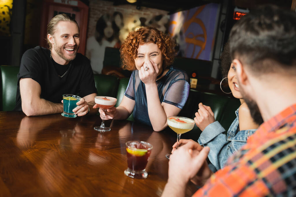 Mujer pelirroja alegre con cóctel sentado cerca de amigos multiétnicos divertirse en el bar  - Foto, Imagen