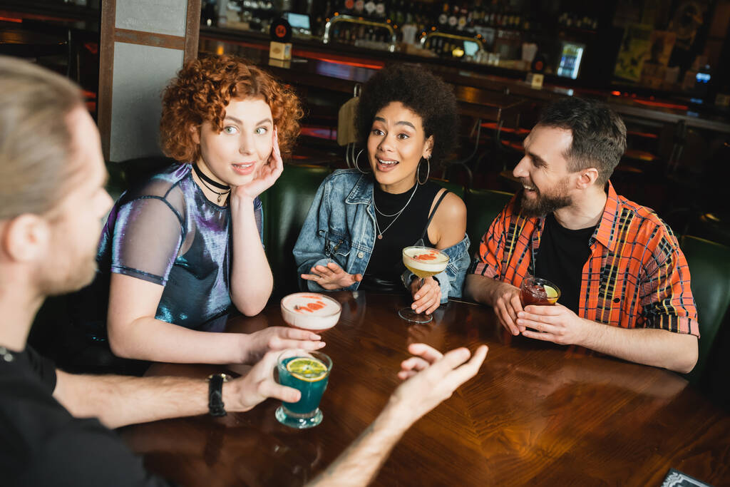 Nadšení multietničtí lidé při pohledu na přítele, zatímco mluví v blízkosti různých koktejlů v baru  - Fotografie, Obrázek