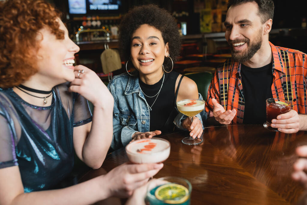 Молодая кудрявая африканская американка держит коктейль рядом с веселыми друзьями в баре  - Фото, изображение