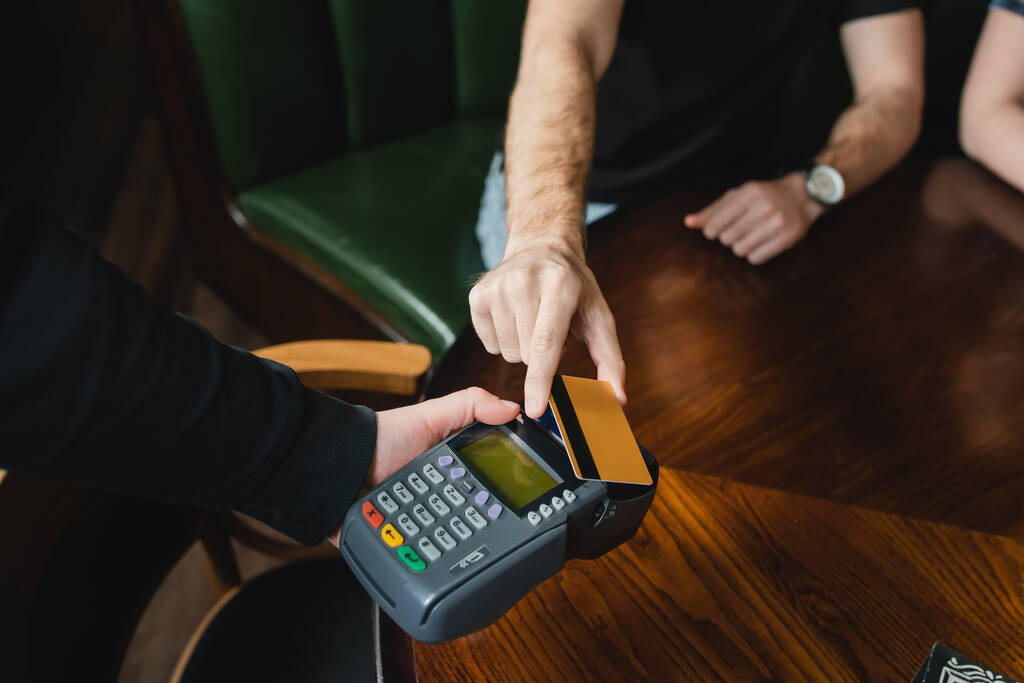 Vista cortada do garçom segurando terminal de pagamento perto do homem com cartão de crédito no bar  - Foto, Imagem
