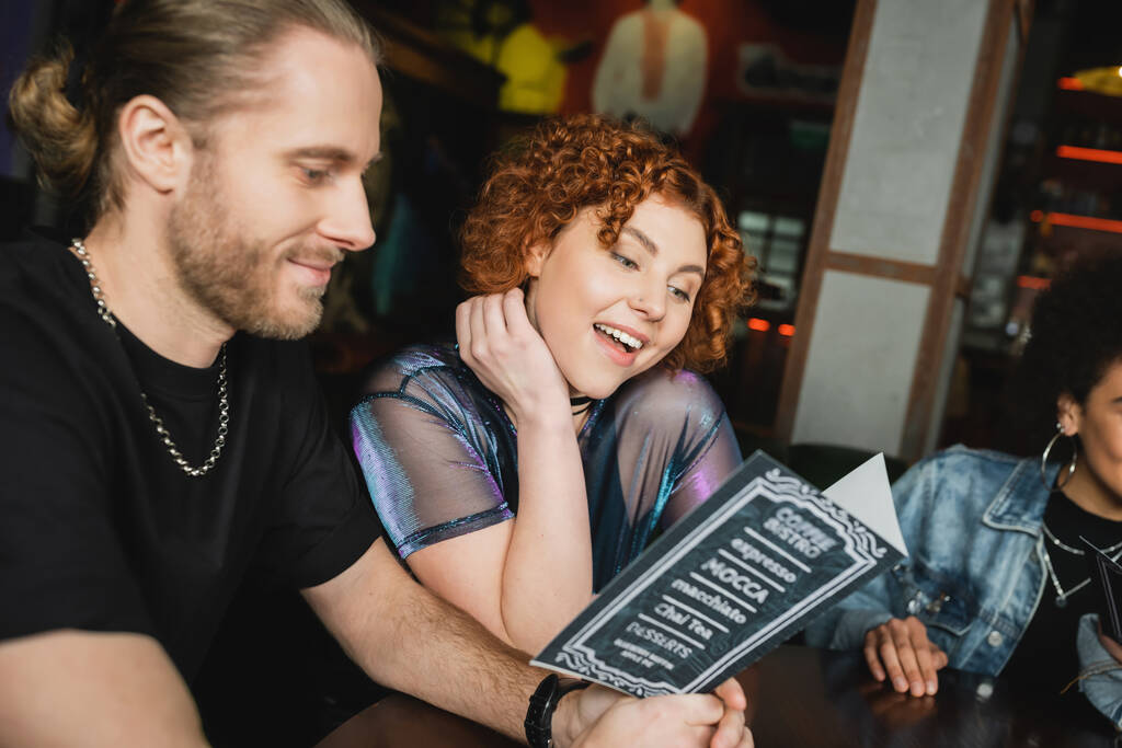 Amigos sonrientes mirando el menú mientras se reúnen en el bar por la noche  - Foto, imagen