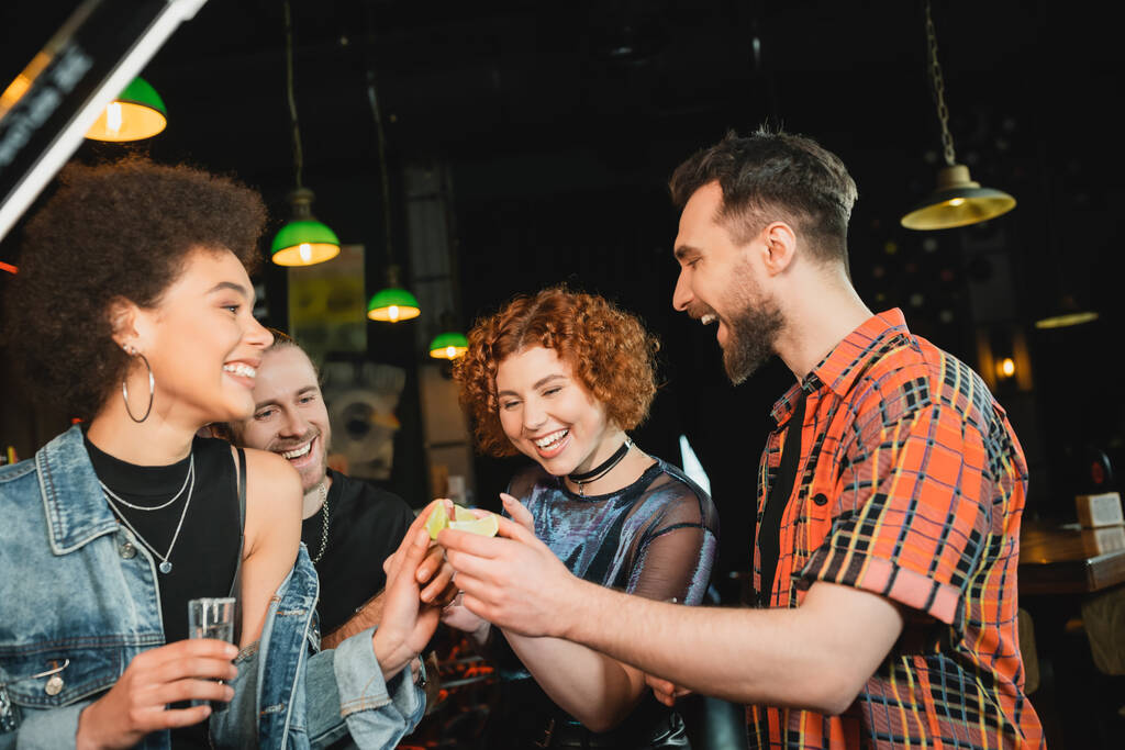 Fröhliche Freunde halten Limetten und Tequila in der Nähe eines afrikanisch-amerikanischen Freundes in der Bar  - Foto, Bild